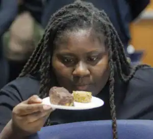 A person examines two pastries on a plate, one chocolate-coated and the other a light-colored treat, with a focused expression. The background is blurred, suggesting a casual setting.