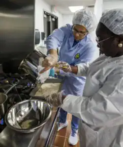 Two people in lab coats and gloves are preparing food in a kitchen, pouring ingredients from a pot into a pan while interacting with each other. A metal bowl is placed nearby on a stove.