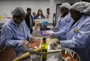Students in lab coats prepare food in a kitchen. Some are cutting ingredients, while others are smiling and observing. Various cooking tools and ingredients are visible on the countertop.
