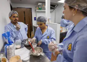 Three people in blue lab coats and hairnets work in a kitchen. One person is mixing ingredients in a bowl, while another observes and holds a spatula. The workspace features various ingredients and utensils.