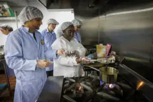 A group of four people in kitchen attire, including hairnets and gloves, are cooking together in a professional kitchen, focused on various cooking tasks. A pot is heating on the stove.