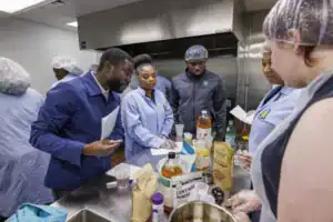 A group of people in a kitchen collaborates at a table filled with various ingredients and containers, discussing recipes and measurements, while wearing hairnets and gloves.