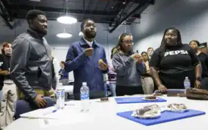 A group of people stands around a table with snacks, engaged in a tasting event. Some hold food items while others watch intently, with bottles of water visible on the table. The setting appears to be a workshop or informal gathering.