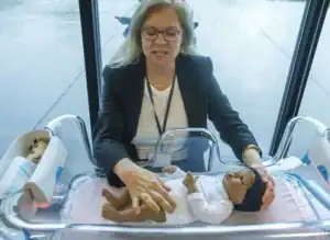 A woman in a blazer gently interacts with a realistic baby doll in a transparent bassinet, demonstrating care techniques. A partially visible toy is in a nearby compartment and rain can be seen through the window.
