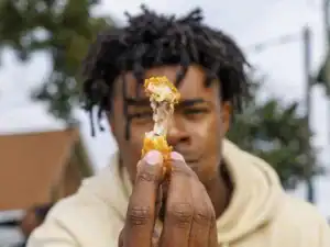 A person holds up a crispy chicken wing while looking intently at the camera. The background features greenery and hints of urban elements.