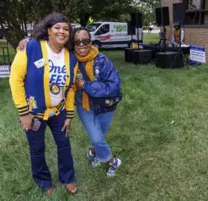 Two women pose together outdoors, smiling and embracing. They wear coordinated blue and yellow outfits representing a college. In the background, there's a U-Haul truck and a DJ setup. Green grass surrounds them.