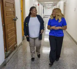 Two women walk side by side down a hallway, engaged in conversation. One wears a white blouse and cardigan, while the other is in a blue polo shirt. The setting is a well-lit corridor with doors on either side.