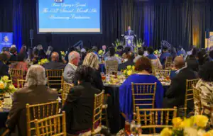 A formal event celebrating the 135th anniversary of the Second Morrill Act. A speaker stands at a podium addressing a seated audience dressed in formal attire, surrounded by decorated tables with floral arrangements.