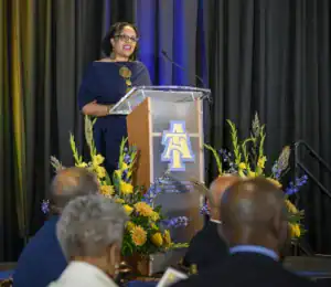 A woman in a blue dress speaks at a podium adorned with flowers. Attendees, seen from behind, are engaged in the presentation. The backdrop features a logo representing a university.