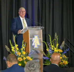 A speaker smiles at a podium decorated with flowers, addressing an audience. The backdrop features the logo of North Carolina A&T State University.
