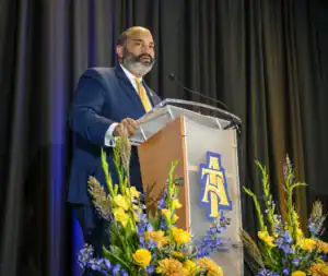 A man with a beard speaks at a podium adorned with flowers, featuring the logo of North Carolina Agricultural and Technical State University. The backdrop is dark with soft lighting.