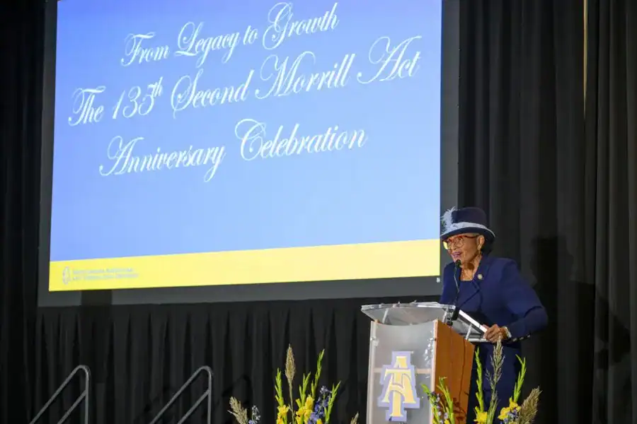 A speaker at a podium addresses an audience during the 135th Anniversary Celebration of the Second Morrill Act, with a large backdrop displaying the event's title.