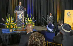 A speaker stands at a podium adorned with flowers, addressing an audience during an event at North Carolina A&T State University. Attendees in formal attire listen attentively.
