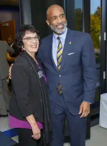 Two smiling individuals pose together in a formal setting. One person wears glasses and a black jacket over a purple top, while the other is in a blue suit with a striped tie.