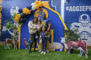 A family poses together in front of a colorful backdrop featuring a small barn and farm animal cutouts. The parents hold their children while smiling, creating a joyful atmosphere.