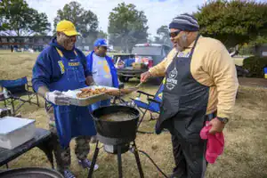 Three men are cooking outdoors at a gathering. One man holds a tray of fried food, while another stirs a pot on a burner. The setting features chairs and a pickup truck in the background.