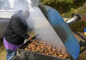 A person using tongs to grill various meats, including chicken and burgers, on a large barbecue smoker surrounded by smoke outdoors.