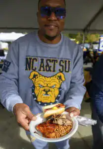 A smiling man in sunglasses holds a plate of barbecue chicken, pulled pork, baked beans, and a hot dog. He wears a sweatshirt featuring the NC A&T logo. A festive outdoor setting is visible in the background.