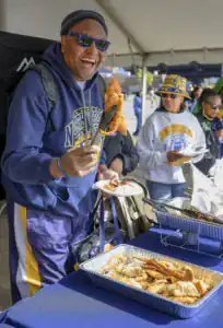 A smiling person in a blue hoodie holds a piece of fried food with tongs, standing at a table filled with different foods. Others are behind him, enjoying the meal in a festive outdoor setting.