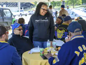 A woman smiles while engaging with a group at a table during a gathering. Attendees are dressed in matching jackets and enjoying food and drinks under a large tent.