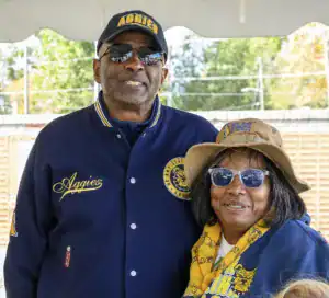 A man and a woman smile together under a tent. The man wears sunglasses and an "Aggies" jacket, while the woman sports a hat and scarf with "Aggies" branding. Lush greenery is visible in the background.
