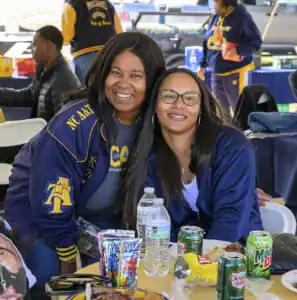 Two women smile at the camera while seated at a table filled with food and drinks. They wear matching jackets and glasses, enjoying a social gathering outdoors. Others are seen in the background.