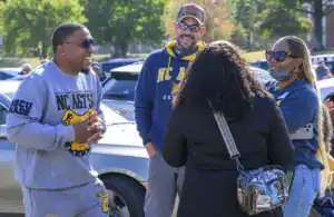 A group of four people chat outdoors, smiling and engaging with each other. Two wear sunglasses, and one is in casual athletic attire. Cars are parked in the background, surrounded by green trees.