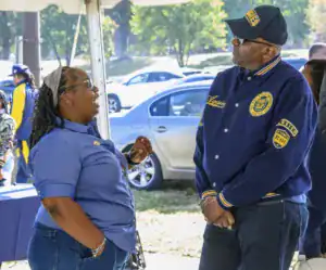 Two people engaged in conversation outdoors. One person is wearing a blue shirt and glasses, while the other is in a navy jacket with yellow accents and a baseball cap. Cars and trees are visible in the background.