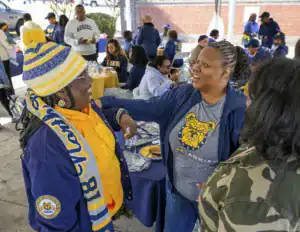 A group of people mingles outdoors, wearing clothing in school colors. Two women engage in conversation, one in a striped hat and jacket, while others enjoy food at nearby tables. The atmosphere is lively and social.