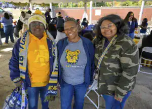 Three women stand together, smiling. One wears an NC A&T hoodie and scarf, another sports a gray NC A&T shirt, and the third has a camouflage jacket. They are at an outdoor event with tables in the background.