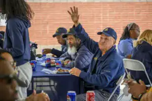 A group of people seated at a table with drinks and food. An elderly man raises his hand while others engage with one another, enjoying a social gathering.