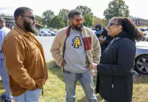 Three people engage in conversation outdoors. One man in a brown corduroy jacket, another in a light sweater with a logo, and a woman in a black jacket. Cars and trees are visible in the background under a bright sky.