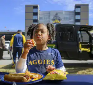 A young girl with curly hair enjoys a hot dog and a plate of food, looking playfully at the camera. A stadium is visible in the background on a sunny day.