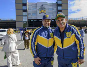 Two men in matching blue and yellow sports outfits smile together in front of a stadium. In the background, others are seen walking and enjoying the atmosphere.