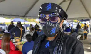 A stylish woman wearing a black mask adorned with bling and large sunglasses, showcasing a blue logo. She's dressed in a black outfit under a tent with people mingling in the background.