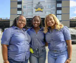 Three smiling individuals in blue button-up shirts pose together outside Truist Stadium, with a car parked nearby and a clear sky in the background.