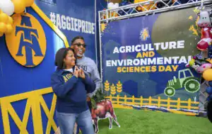 A woman and man pose happily at an event celebrating Agriculture and Environmental Sciences. The background features a colorful banner and decorations, including a cow and tractor.