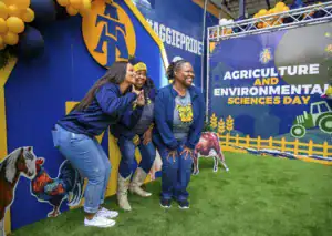 Three women pose playfully in front of a vibrant backdrop celebrating Agriculture and Environmental Sciences Day, with playful animal cutouts, colorful balloons, and the university logo prominently displayed.