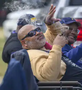A man in sunglasses enjoys a cigar while reclining, exhaling smoke. Another smiling man sits nearby, both engaged in a relaxed outdoor setting.