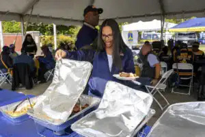 A woman in a navy jacket serves herself food from a tray at a gathering under a tent. A group sits at tables in the background, enjoying the event.
