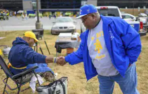 Two men shake hands outdoors, one seated and wearing a yellow cap and a dark hoodie, the other standing in a blue jacket and gray sweatshirt. A parking lot and gathering crowd are visible in the background.