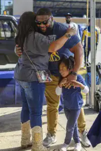 A joyful reunion takes place between a woman, a man, and a young girl. The woman embraces the man while the girl smiles and holds onto both of them. They are outdoors near a table and vehicles in the background.