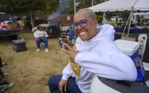 A smiling woman with short, platinum hair and large glasses makes a peace sign. She wears a white hoodie with a university logo while sitting outdoors at a social gathering with people and food in the background.