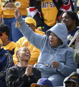 Fans celebrate in a stadium. One woman in a gray hoodie joyfully raises a cheerleading pom-pom, while another woman in a black sweatshirt smiles and claps. People around them wear matching team colors.