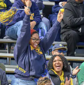 A jubilant fan cheers with a pom-pom while another woman laughs joyfully beside her, both in casual team colors, celebrating at a sporting event in a bleacher setting.