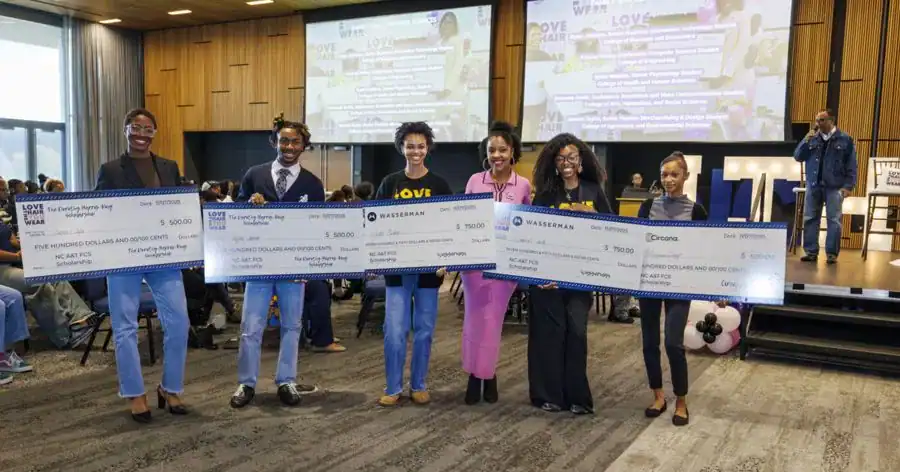 Participants hold large ceremonial checks at an event, celebrating scholarship awards. The background features a presentation screen and an audience.