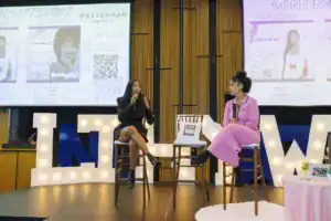 A speaker in a black outfit shares insights while seated on a stool during a live interview. Another participant in a pink dress listens attentively, with signage and a colorful backdrop in the venue.