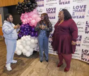 Three individuals interact at an event, standing in front of a backdrop with colorful balloons and text promoting self-love and hair care. One wears a gray sweatsuit, another a black jacket, and the third a maroon outfit.