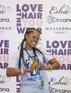 A smiling person with braided hair poses in a gray shirt that says "FIRST GEN." They are making a hand gesture in front of a backdrop featuring text about loving hair and various sponsor logos.