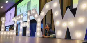 A child in sunglasses lies beside large, illuminated letters spelling "LEHYW" on a stage. Colorful screens are visible in the background, displaying event information.